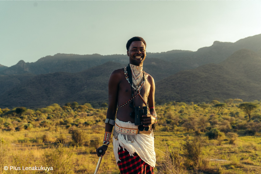 Un homme kenyan porte des vêtements traditionnels, debout devant une belle vue. Il utilise une canne blanche.